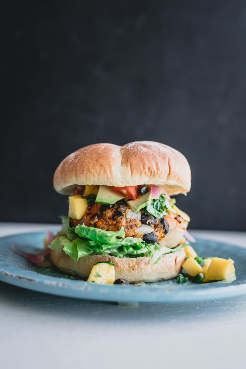 A veggie burger piled high with lettuce, avocado, mango, onion, and tomato on a blue plate.